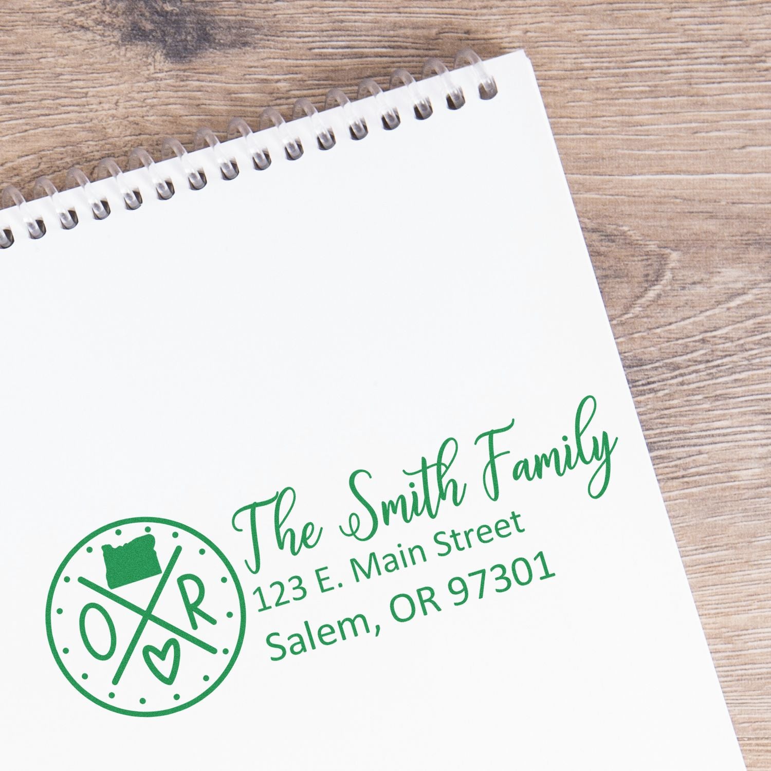 Slim Oregon Customized Pre-Inked Address Stamp on a notepad, displaying The Smith Family, 123 E. Main Street, Salem, OR 97301 in green ink, with a decorative Oregon state symbol.