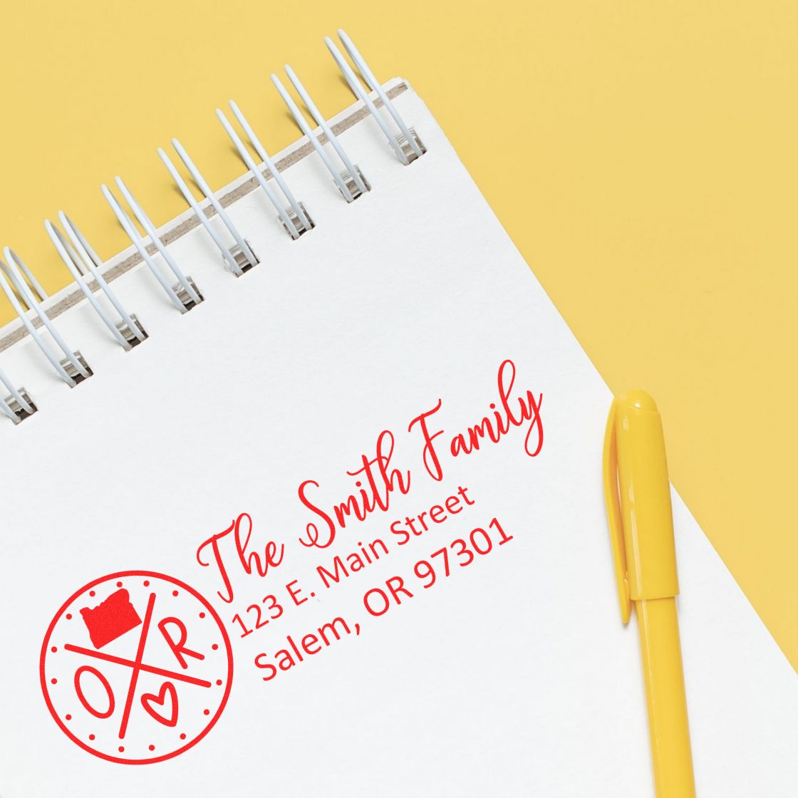 Slim Oregon Customized Pre-Inked Address Stamp in red ink on white paper, displaying The Smith Family, 123 E. Main Street, Salem, OR 97301 with a yellow pen on a yellow background.