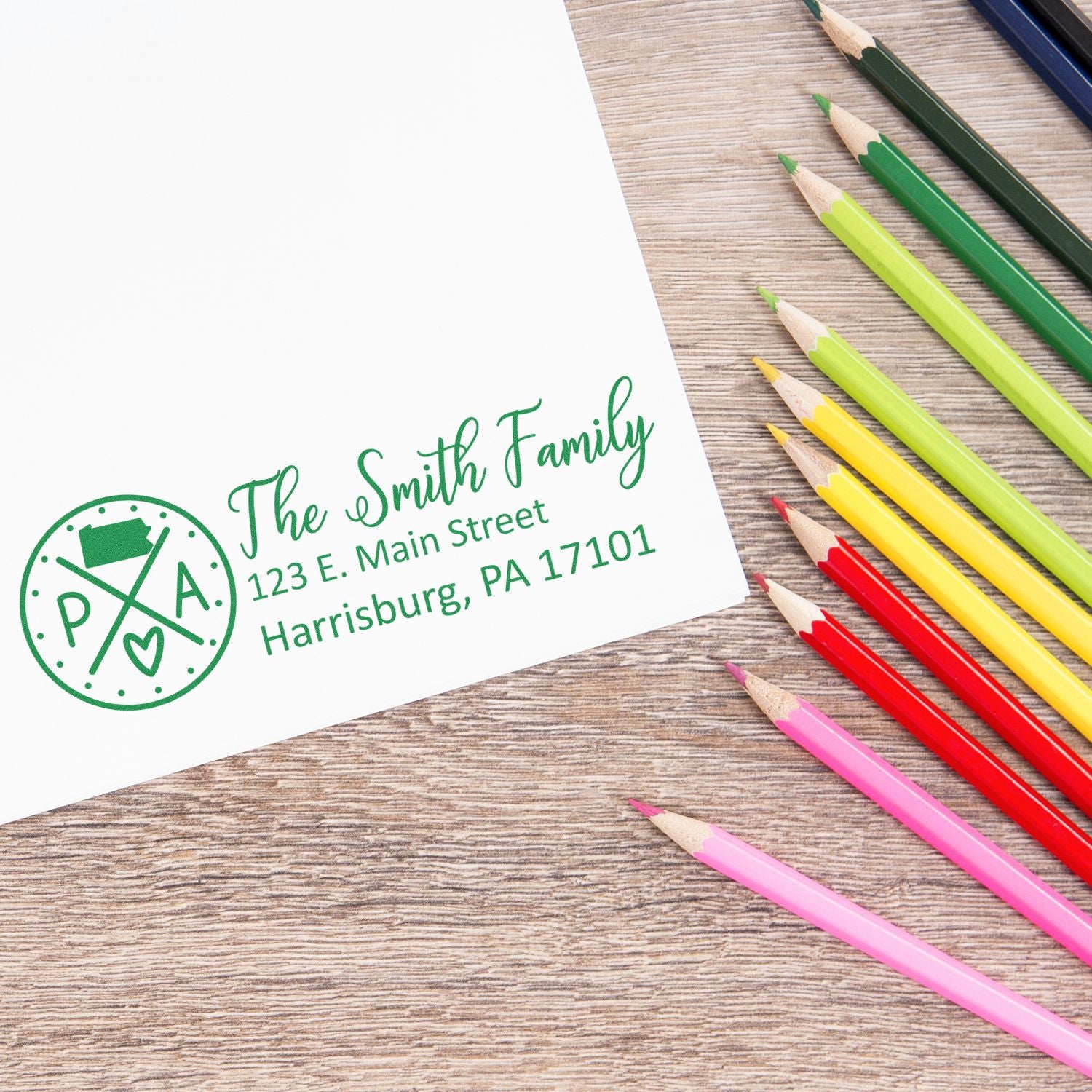 Slim Pennsylvania Customized Pre-Inked Address Stamp on white paper with colorful pencils. Displays The Smith Family, 123 E. Main Street, Harrisburg, PA 17101 in green ink.