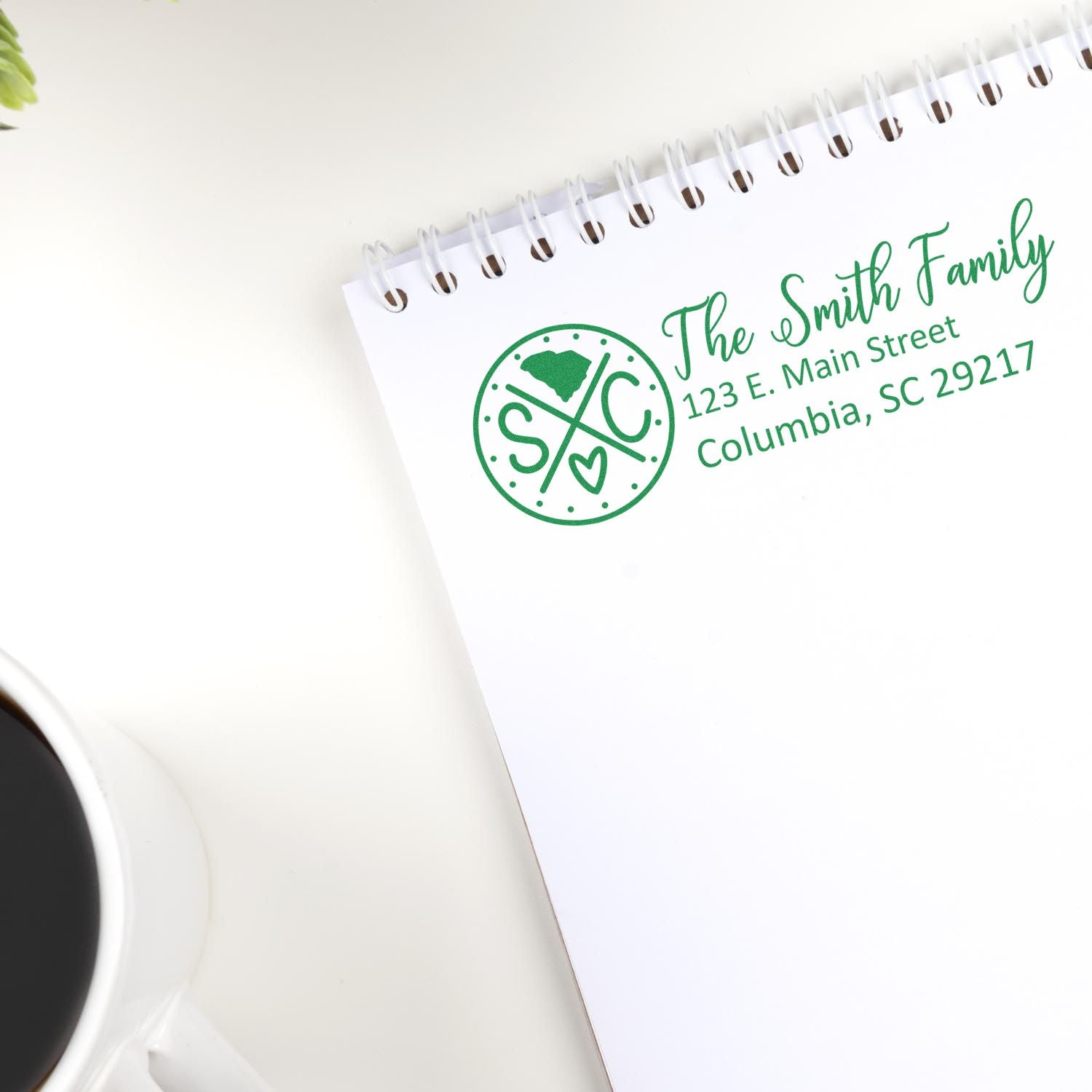 South Carolina State Pride Customized Address Stamp on a notepad with The Smith Family and address in green ink. A coffee cup and plant are nearby on a white surface.