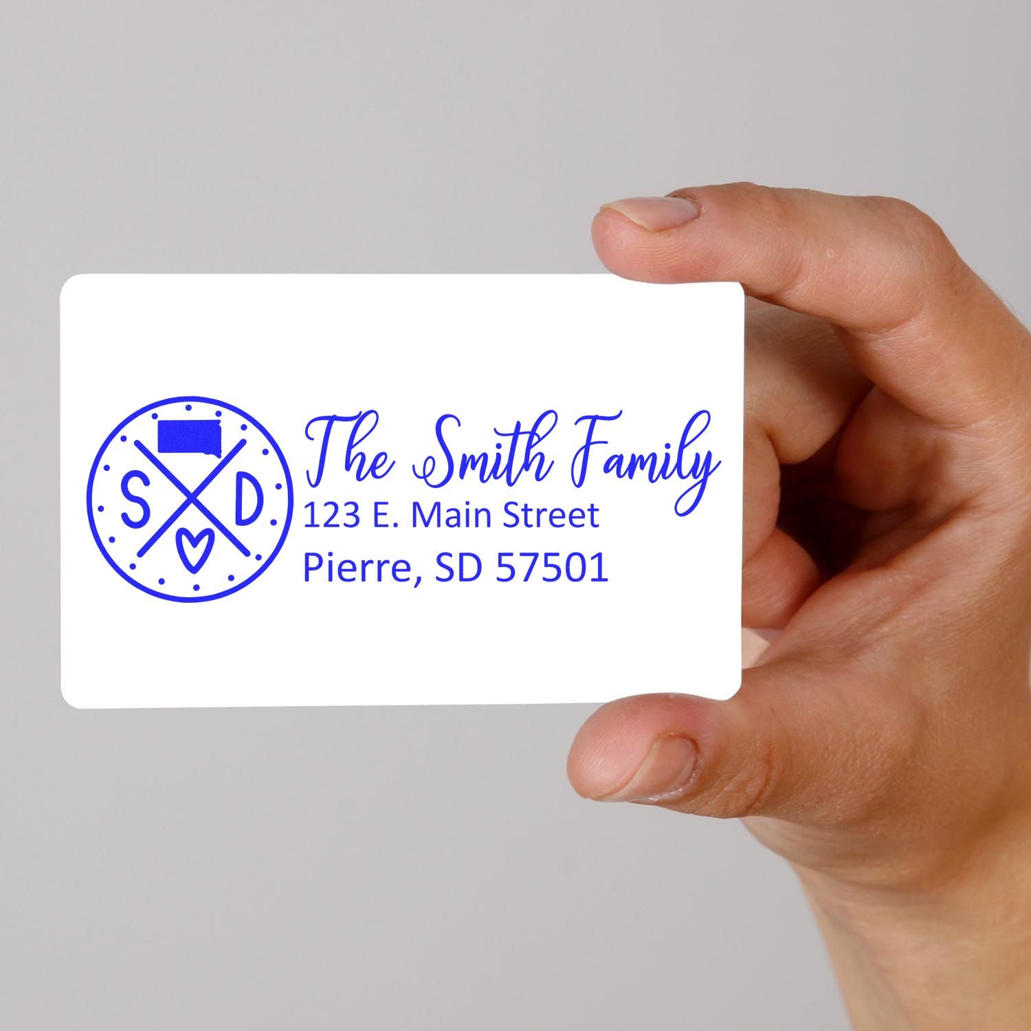 Hand holding a card with a blue Slim South Dakota Customized Pre-Inked Address Stamp design, featuring The Smith Family and an address in Pierre, SD.