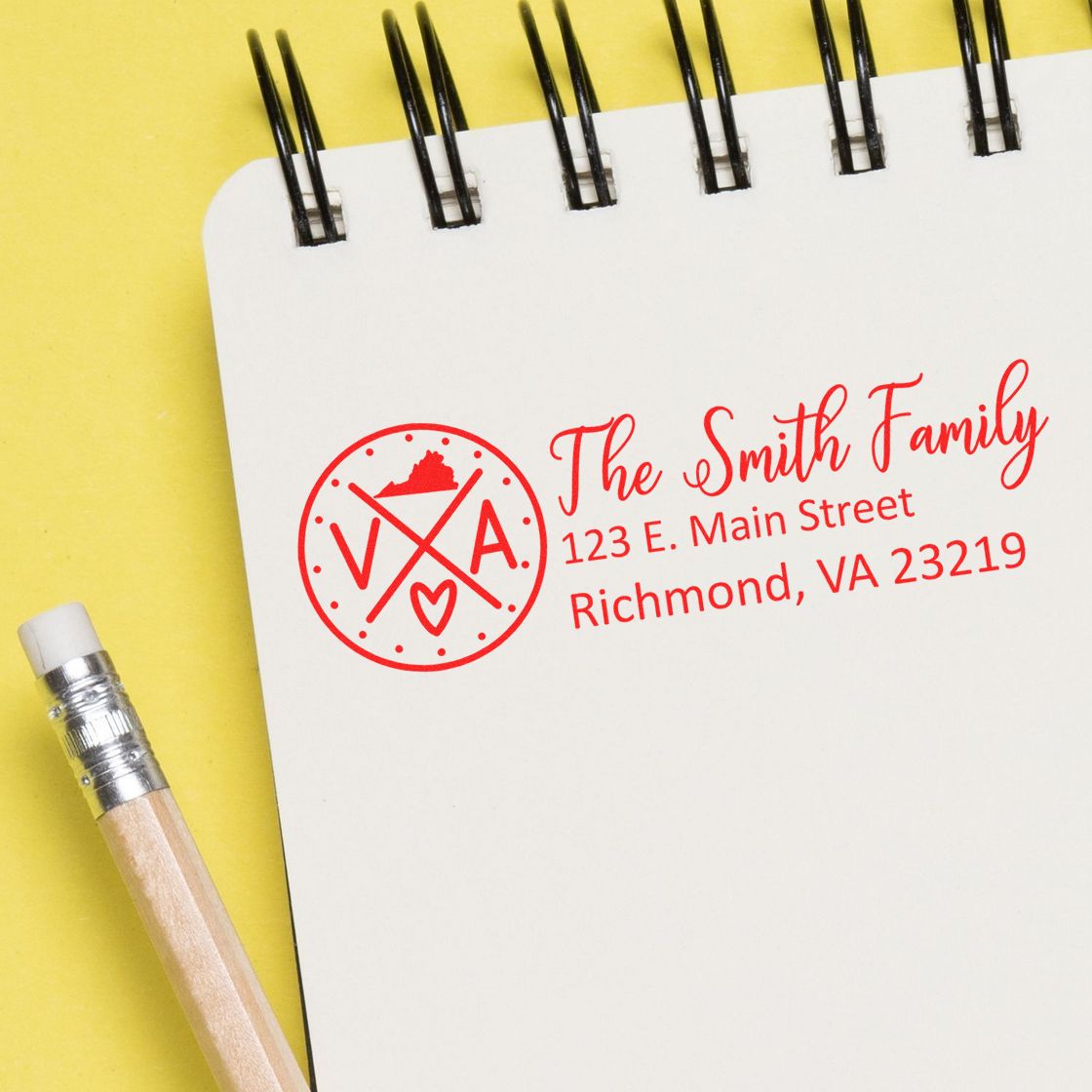 Slim Virginia Customized Pre-Inked Address Stamp on a notepad, displaying The Smith Family, 123 E. Main Street, Richmond, VA 23219 in red ink, next to a pencil on a yellow background.