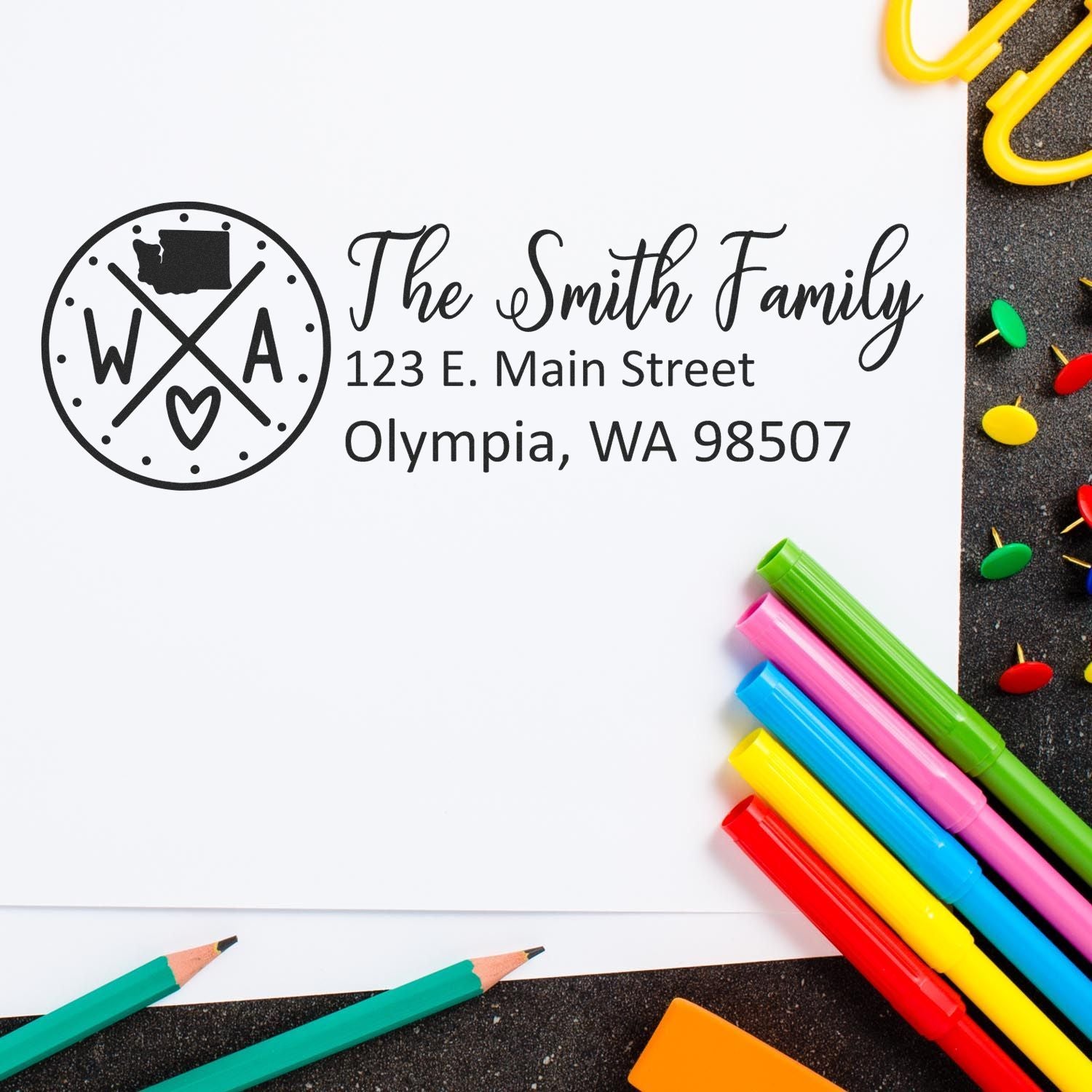 Washington State Pride Customized Address Stamp on white paper with colorful pens and paperclips. The stamp features a circular design with WA and a heart, alongside a sample address for Olympia, WA.