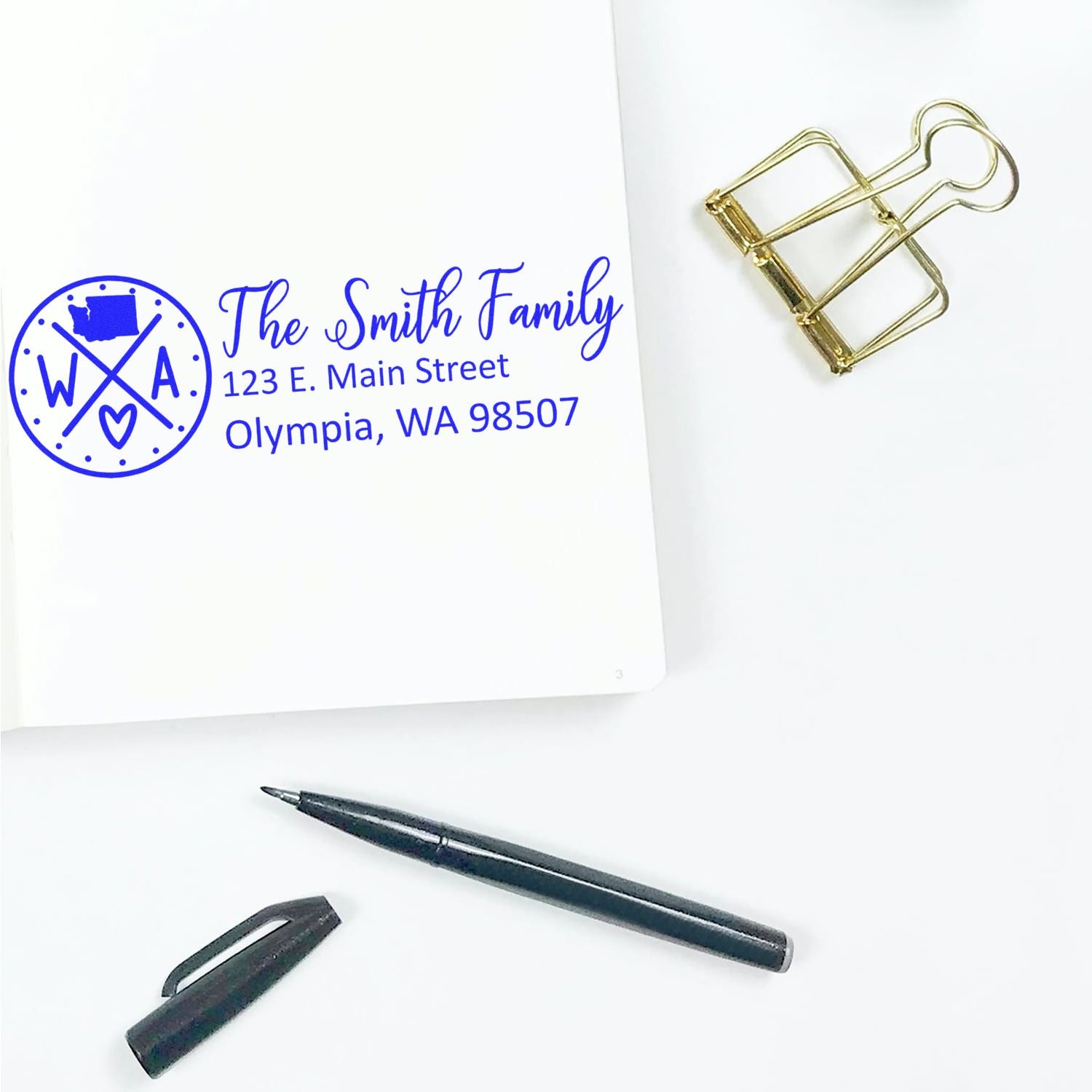 Washington State Pride Customized Address Stamp on white paper, featuring a blue design with state outline and heart. Nearby are a black pen, cap, and gold binder clip on a white surface.