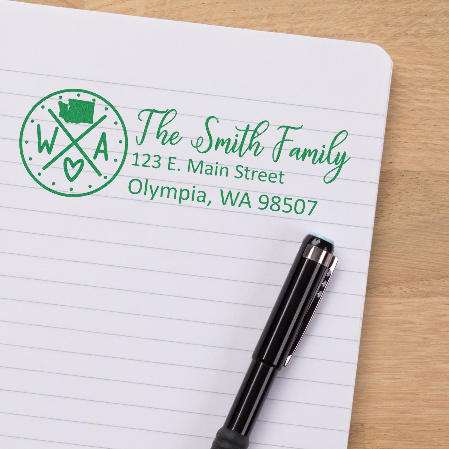 Washington State Pride Customized Address Stamp on paper with The Smith Family, 123 E. Main Street, Olympia, WA 98507 in green ink, next to a black pen on a wooden surface.