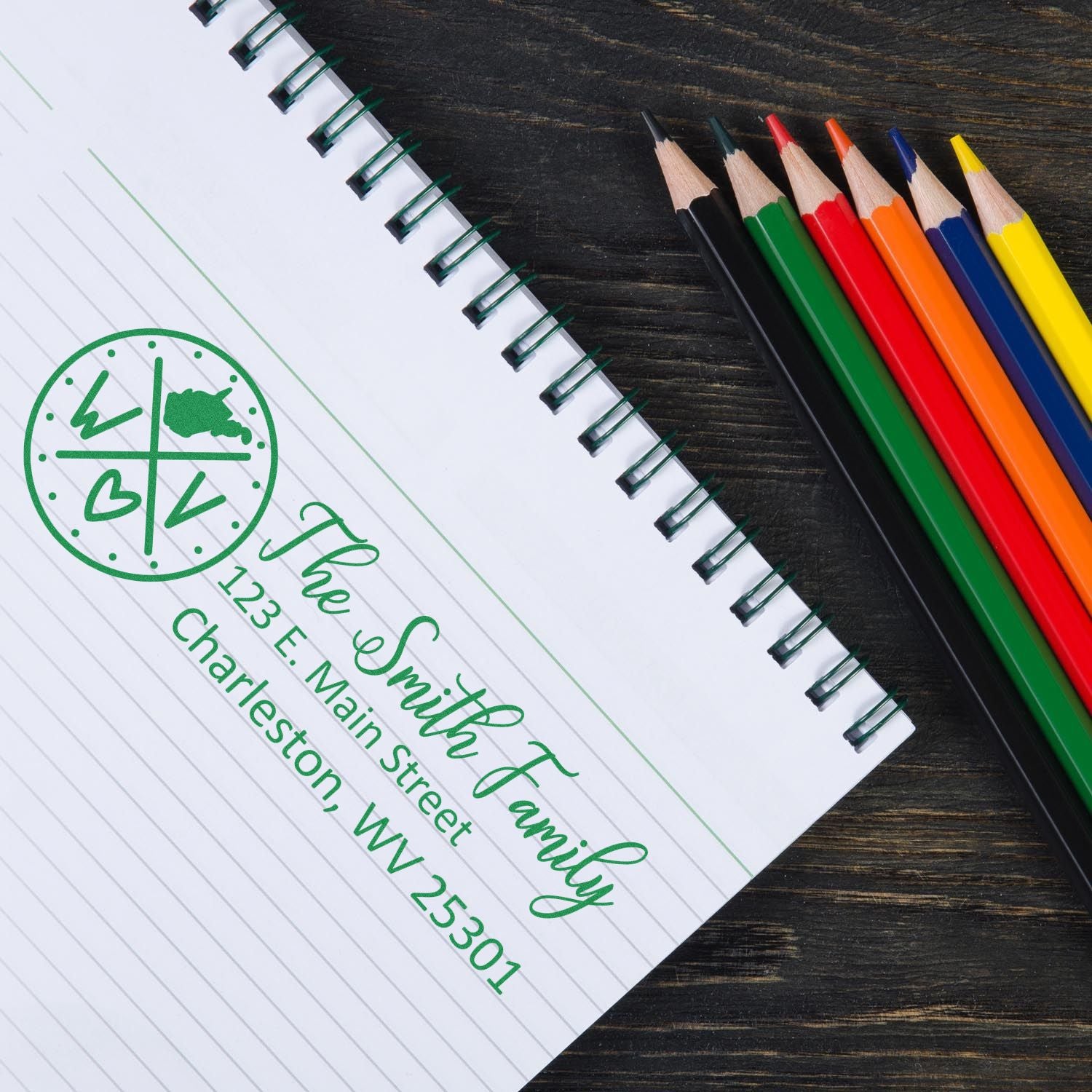 A notebook with a green Self-Inking State Love of West Virginia Custom Address Stamp imprint, showing a family name and address, next to colorful pencils on a dark wooden surface.