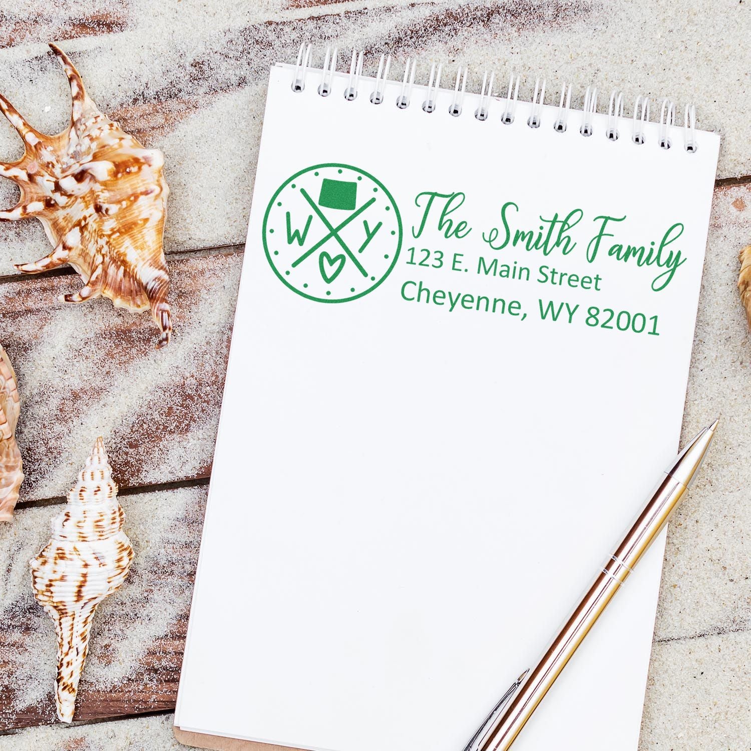 Notebook on sandy surface with seashells, featuring a green PSI Pre-Inked Wyoming State Pride Customized Address Stamp design, displaying The Smith Family and a Cheyenne, WY address.