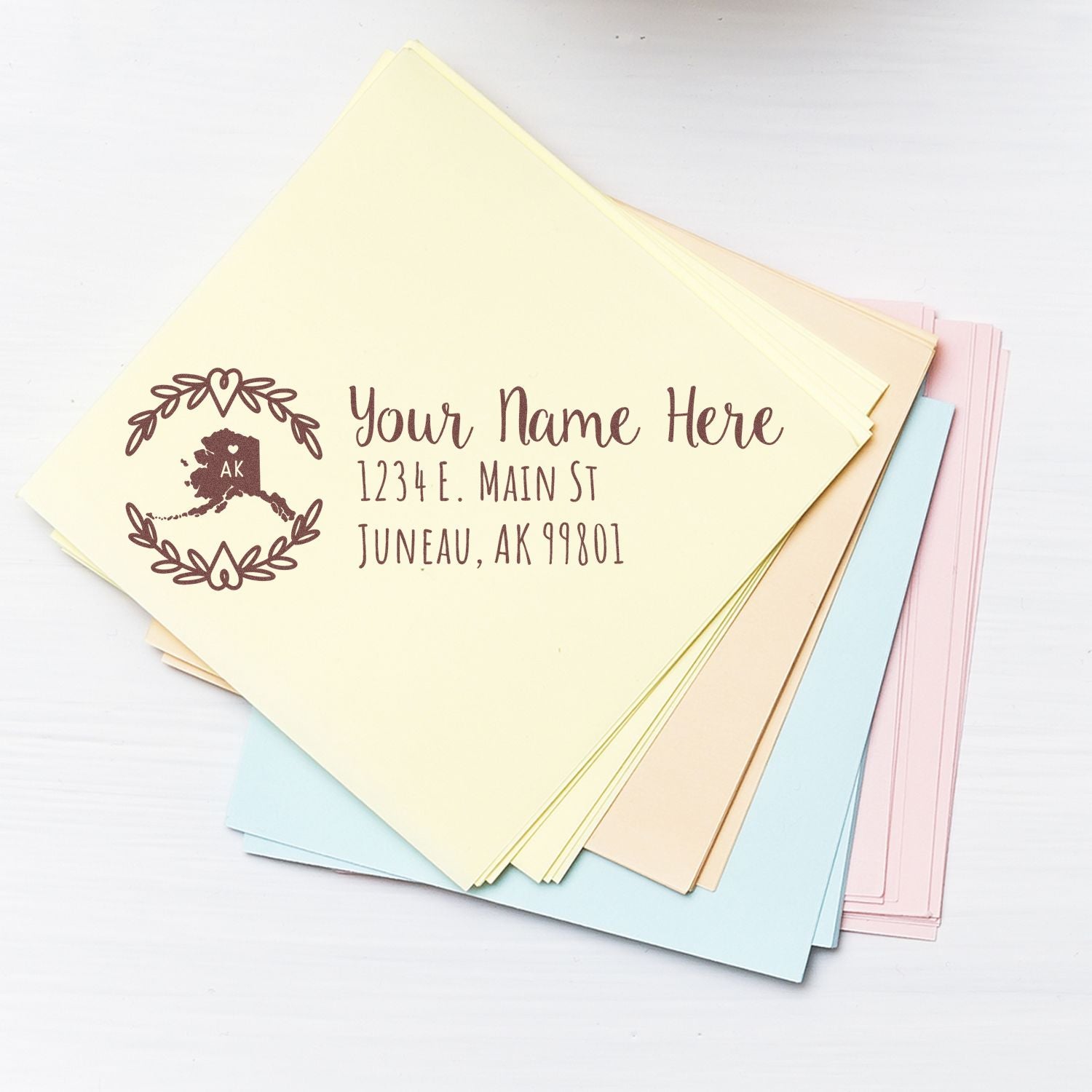 Stack of pastel envelopes with a PSI Pre-Inked Alaska State Customized Address Stamp featuring a floral design and sample address in Juneau, AK.