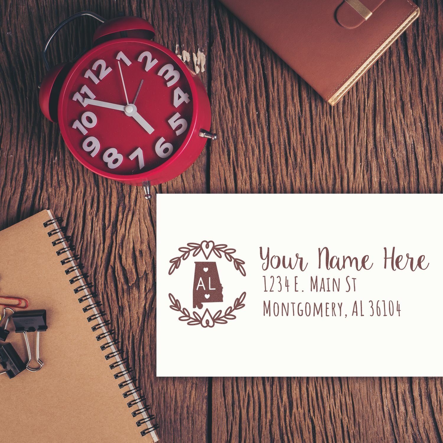 Self-Inking State Wreath of Alabama Address Stamp on an envelope, surrounded by a red clock, notebook, and leather journal on a wooden desk.