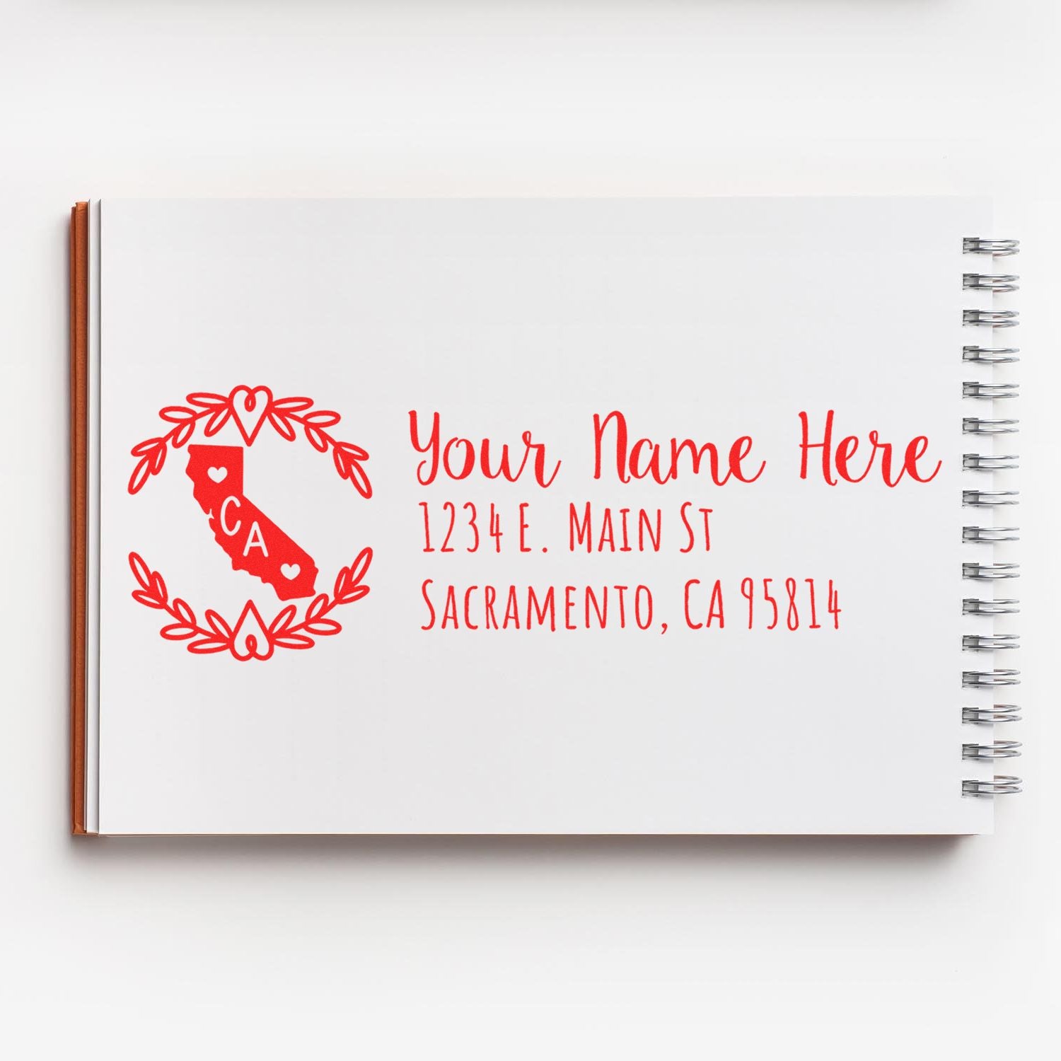 Self-Inking State Wreath of California Address Stamp on a notebook, featuring a red California map with a heart and wreath design, alongside customizable address text in elegant red font.