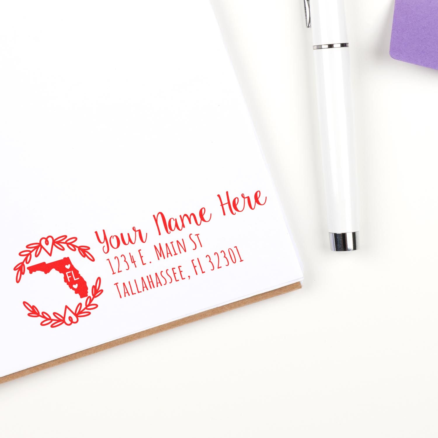 Florida State Custom Return Address Stamp in red ink on white paper, featuring a Florida outline with address details. A pen and purple envelope are nearby.