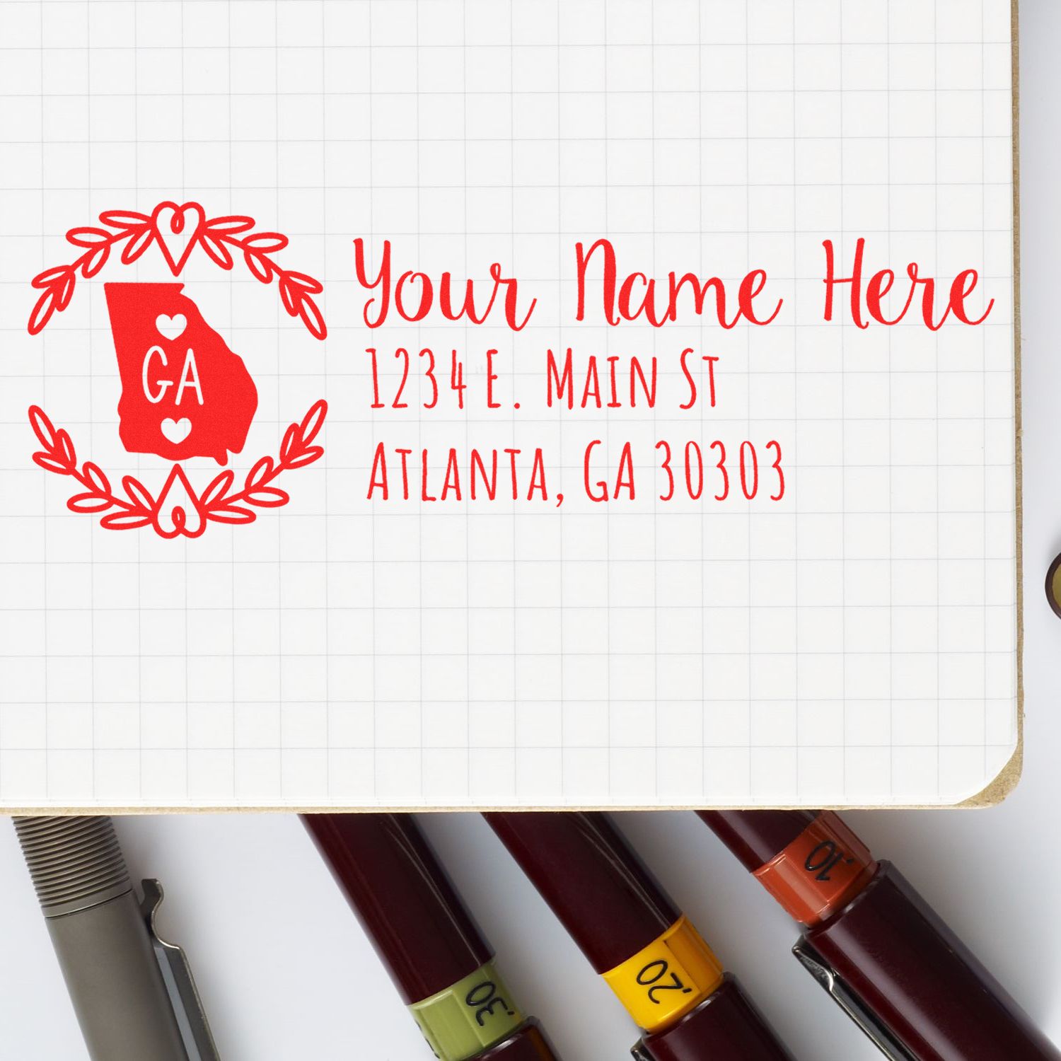 Georgia State Custom Return Address Stamp in red ink on a notebook page, featuring a heart-accented state outline and sample address. Pens are visible at the bottom.