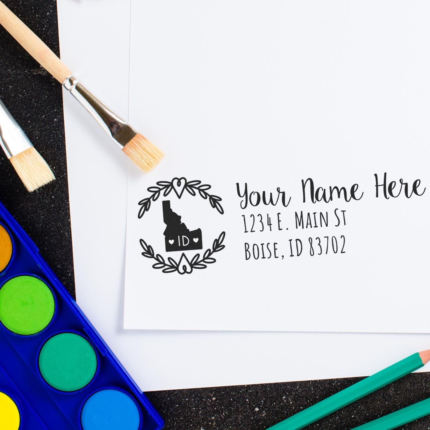 Idaho State Custom Return Address Stamp on white paper, featuring a decorative Idaho state outline with space for personalized name and address. Surrounded by art supplies on a black surface.