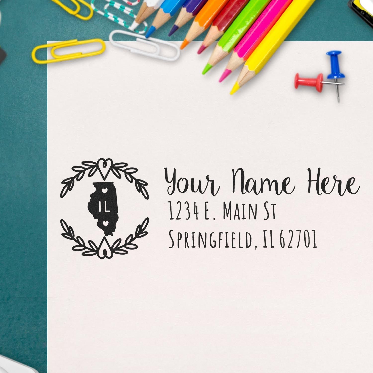 Slim Illinois Personalized Pre-Inked Address Stamp on paper with colorful pencils, paperclips, and push pins. Features a heart design and Illinois outline, showcasing a sample address in elegant script.