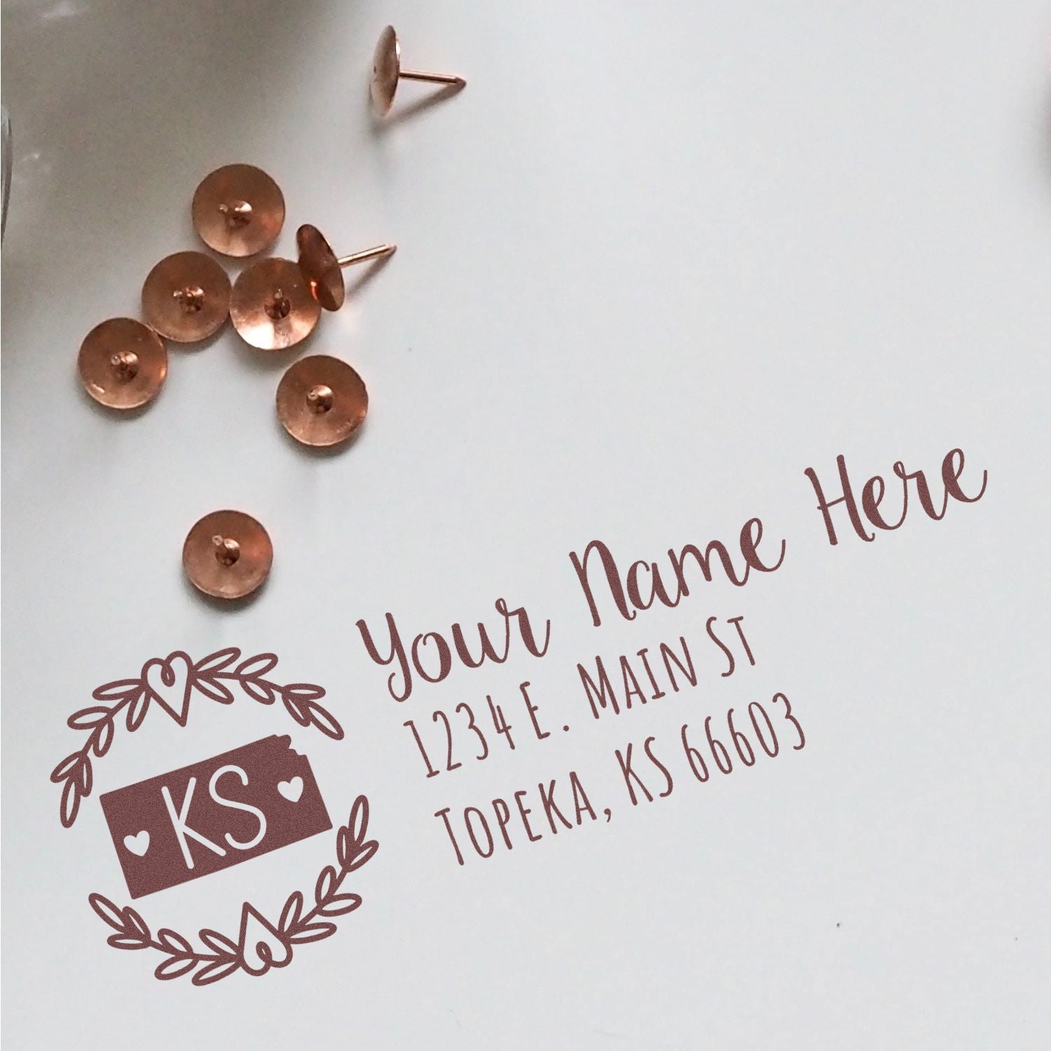 Self-Inking State Wreath of Kansas Address Stamp on white paper with sample text, surrounded by copper push pins. Elegant design featuring KS in a wreath.