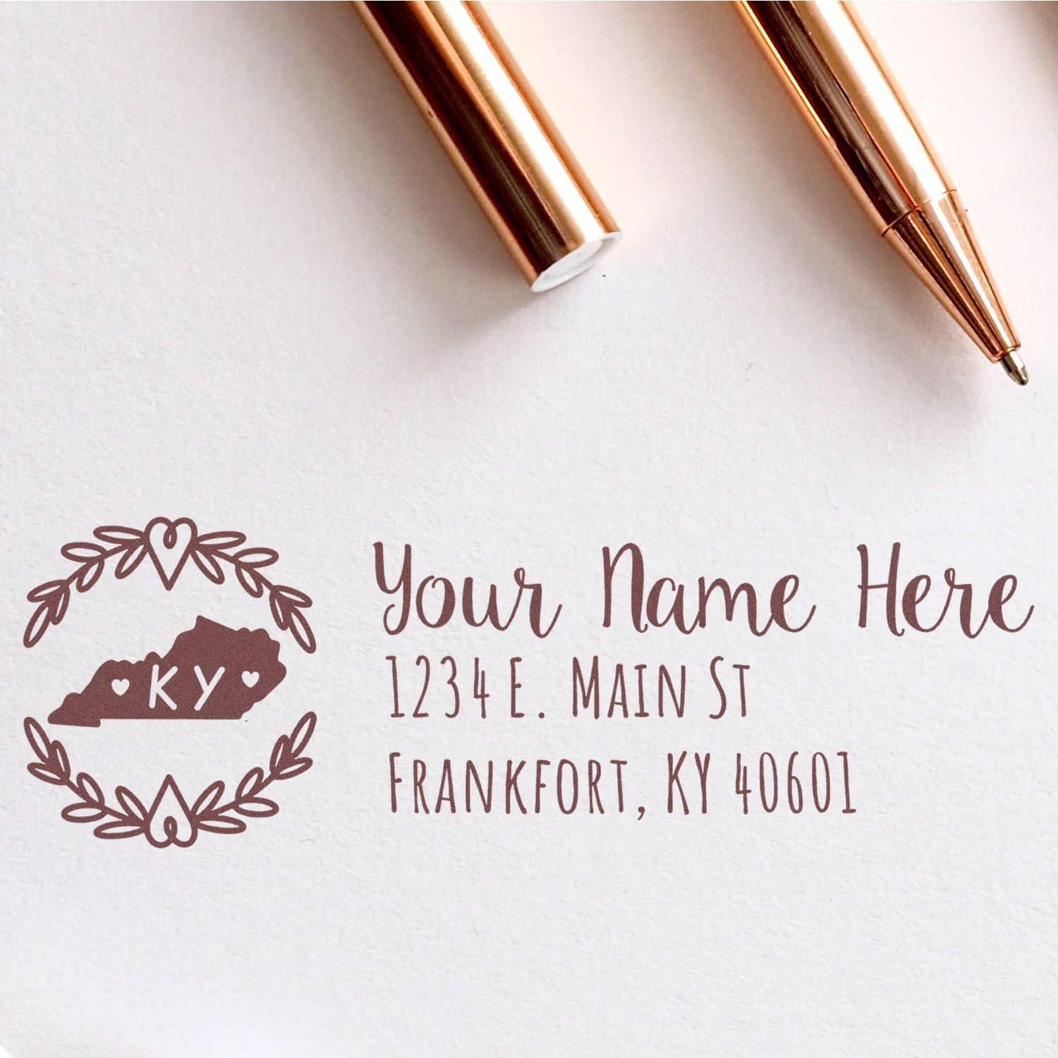 PSI Pre-Inked Kentucky State Customized Address Stamp on white paper with a Kentucky outline, hearts, and sample address in elegant script. Two rose gold pens are placed nearby.