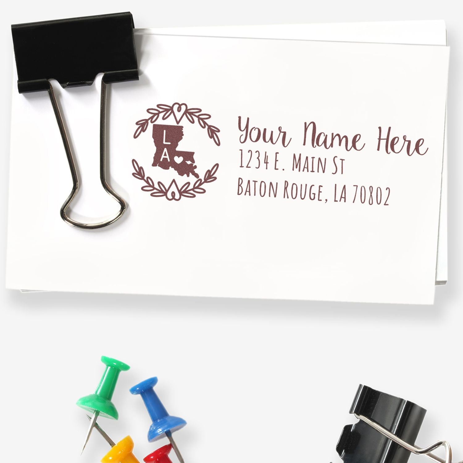 Slim Louisiana Personalized Pre-Inked Address Stamp on white envelope with a decorative state outline and text. Surrounded by colorful push pins and a black binder clip.