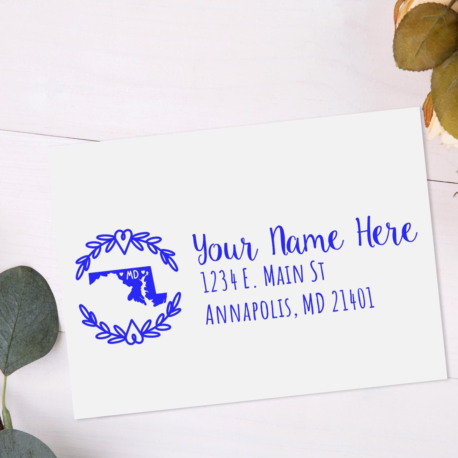 PSI Pre-Inked Maryland State Customized Address Stamp on a white envelope with blue ink, featuring a Maryland map design and sample address. Surrounded by eucalyptus leaves on a light wood surface.