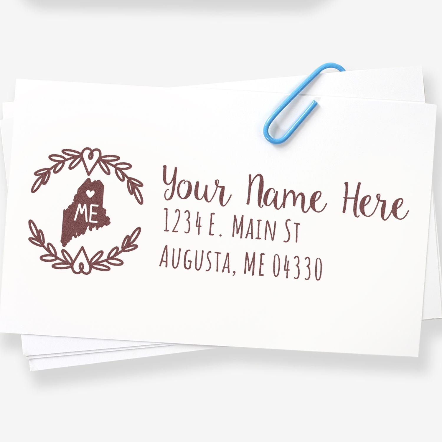 Image of the Slim Maine Personalized Pre-Inked Address Stamp on a stack of envelopes, featuring a heart design and sample address in elegant script, secured with a blue paperclip.