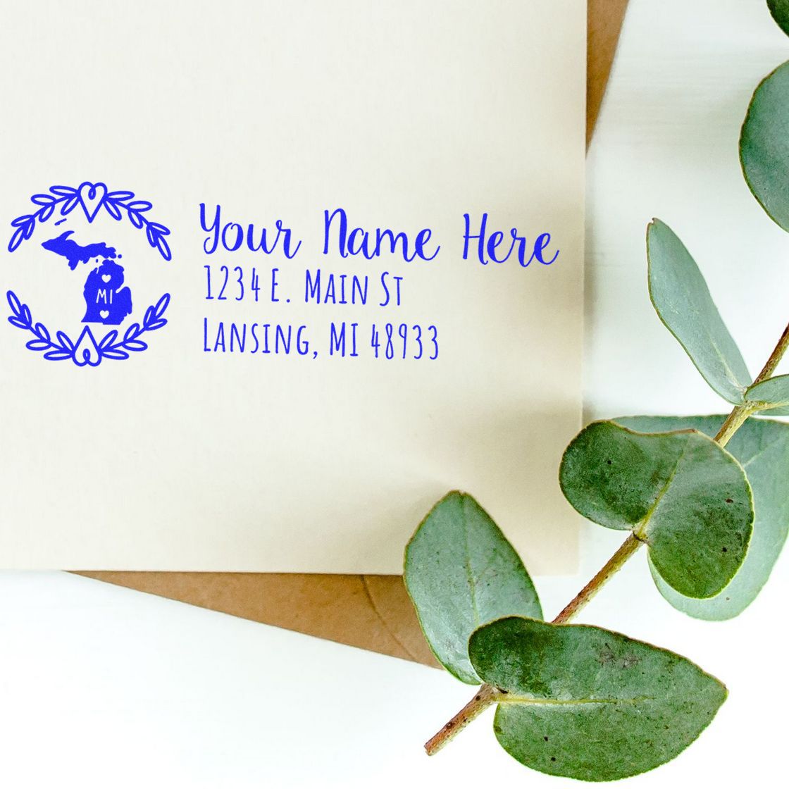 Michigan State Custom Return Address Stamp on an envelope, featuring a blue design with the state outline and decorative leaves, placed next to green eucalyptus branches on a white surface.