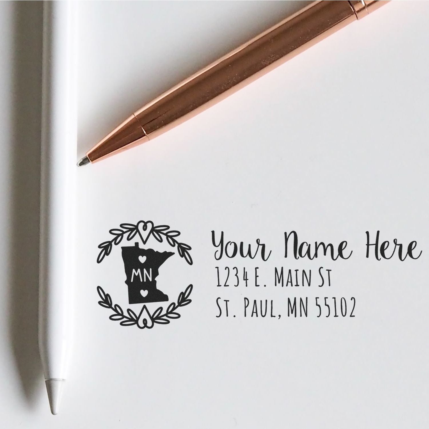 Minnesota State Custom Return Address Stamp on white paper with a rose gold pen and a white pen nearby, featuring a heart design and placeholder text for name and address.