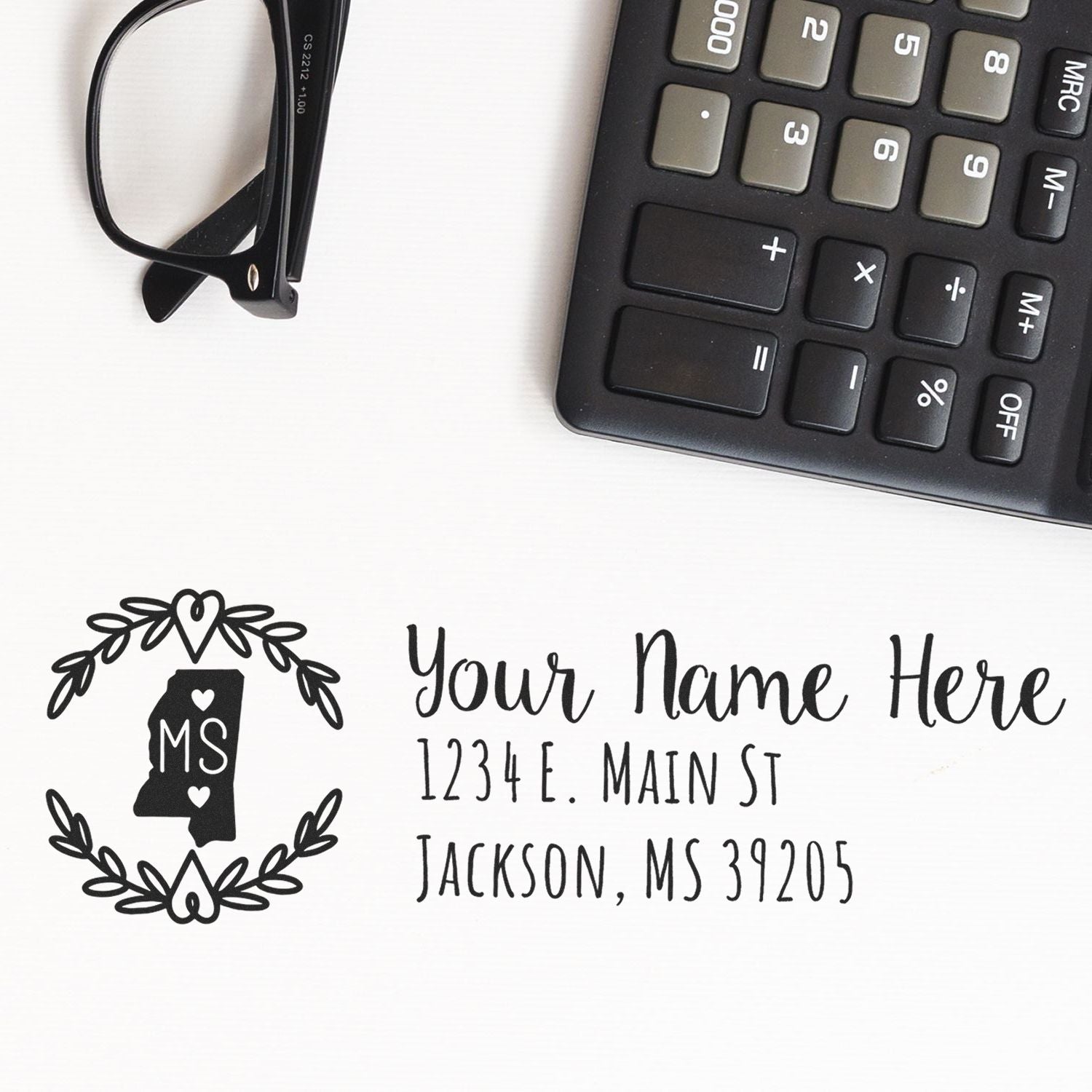 Mississippi State Custom Return Address Stamp on white paper, featuring a decorative state outline with MS and sample address text. Nearby are black glasses and a calculator.