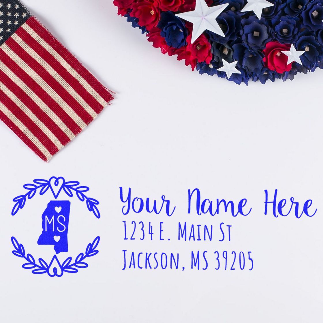 Self-Inking State Wreath of Mississippi Address Stamp on white paper with blue text, featuring a heart and state outline design. Nearby are red, white, and blue decorations with stars.