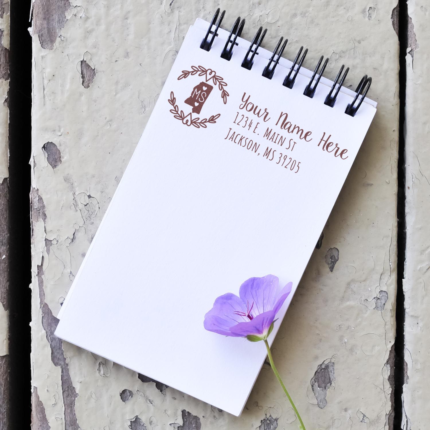 Notepad with PSI Pre-Inked Mississippi State Customized Address Stamp, featuring a floral design and sample address. A purple flower rests on the notepad, placed on a rustic wooden surface.