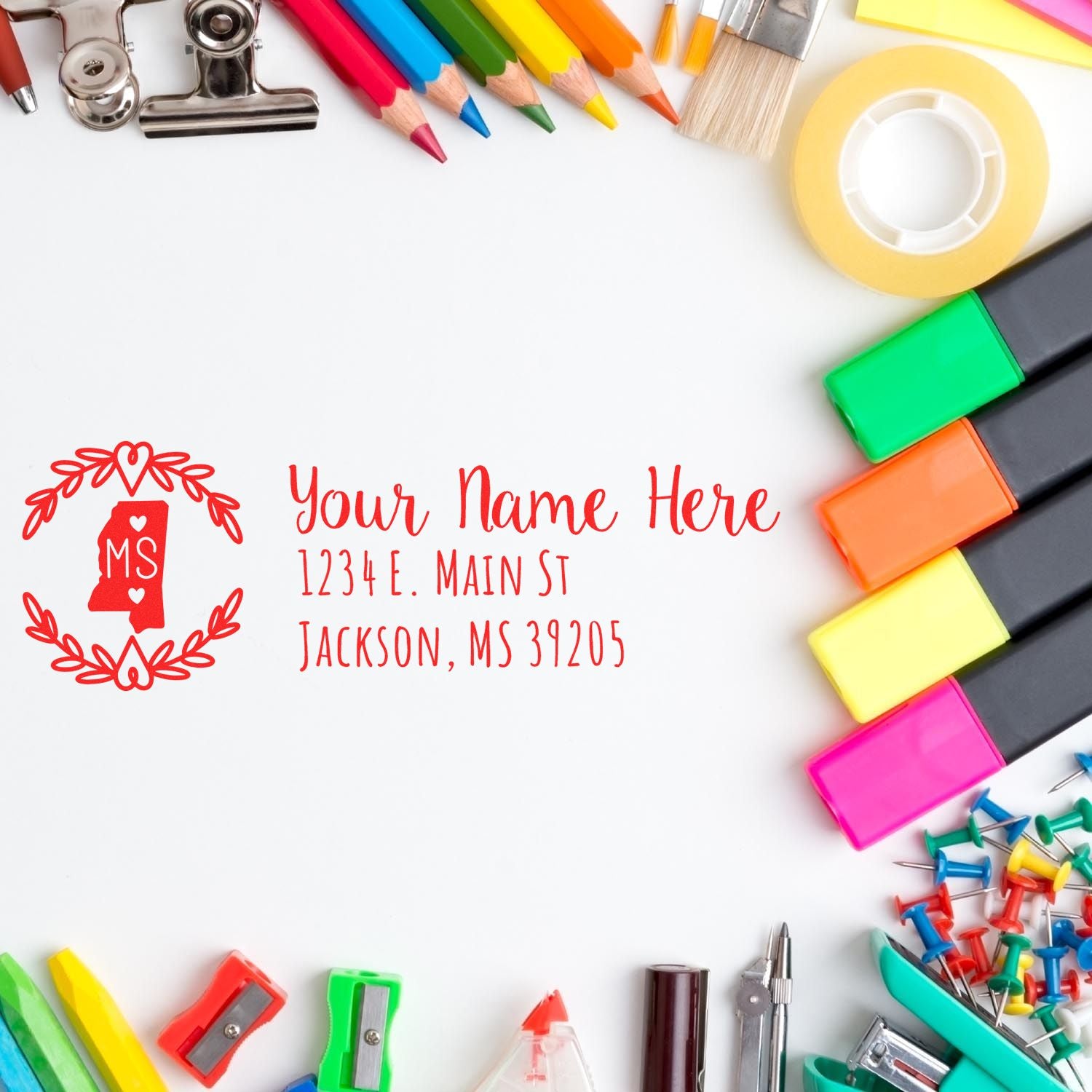 A colorful desk with stationery surrounds a stamped envelope featuring the Slim Mississippi Personalized Pre-Inked Address Stamp, displaying a custom name and address in red ink.