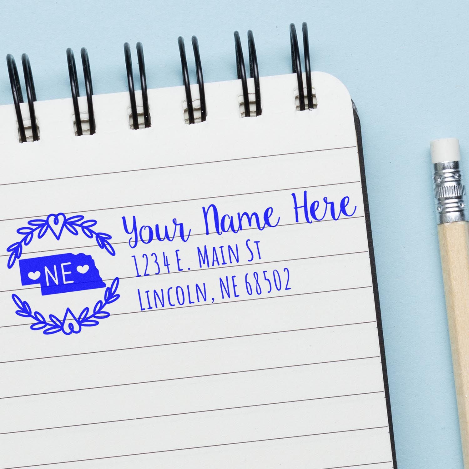 Slim Nebraska Personalized Pre-Inked Address Stamp on a notebook page, featuring a blue design with a state outline and floral border, next to a pencil on a light blue background.