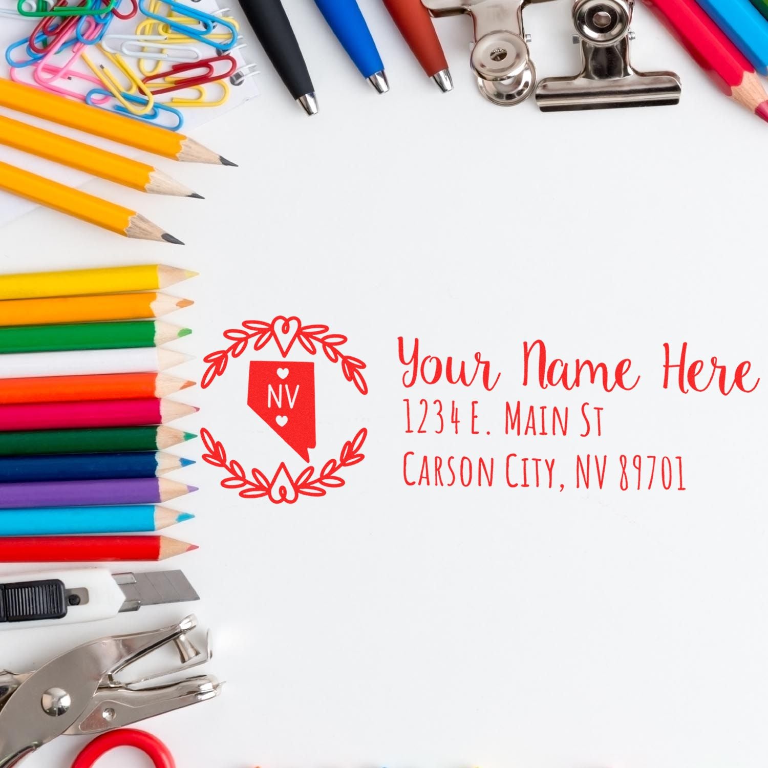 A colorful desk with pencils, paperclips, and a Self-Inking State Wreath of Nevada Address Stamp imprint showing Your Name Here and an address in red ink on white paper.