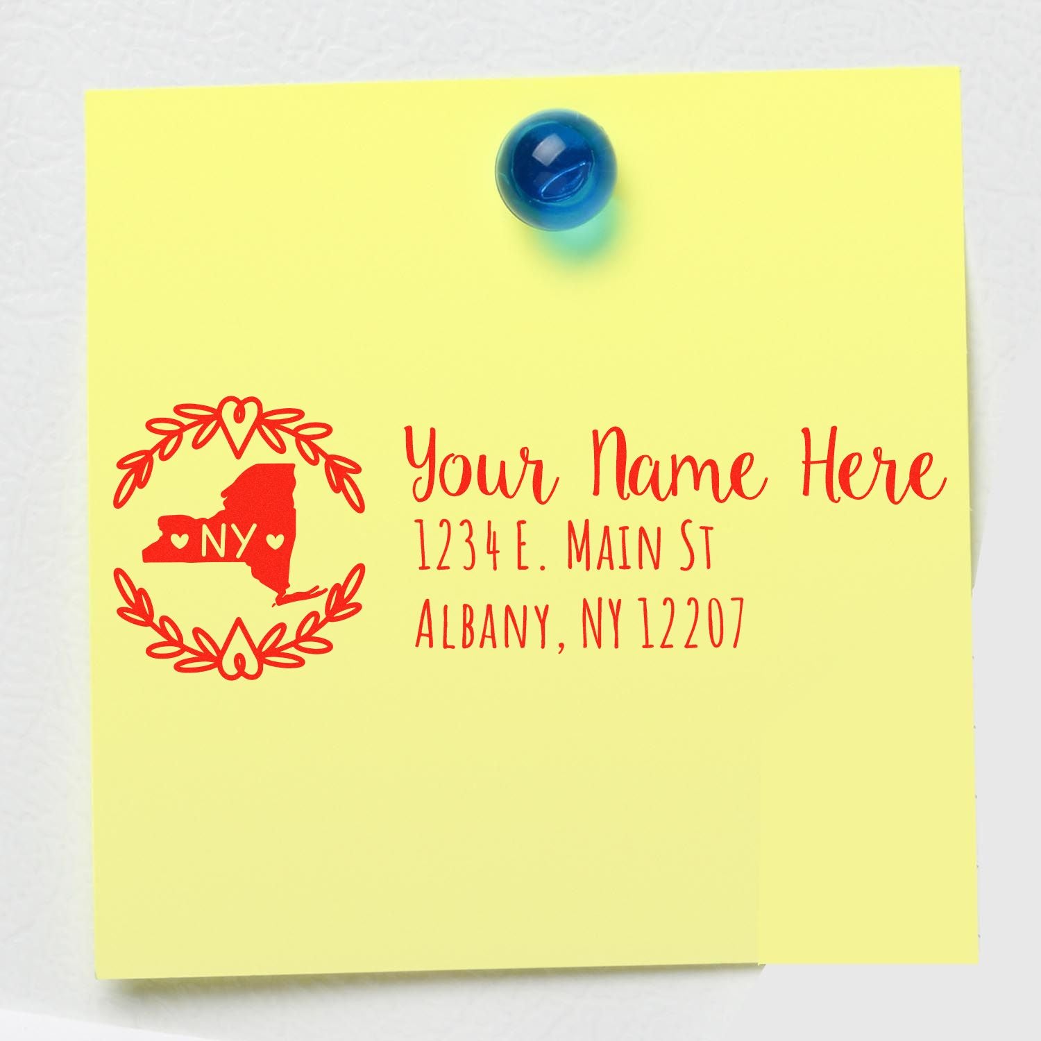 Yellow note with red text stamped using the Slim New York Personalized Pre-Inked Address Stamp, featuring a New York state outline and address details, pinned with a blue magnet.