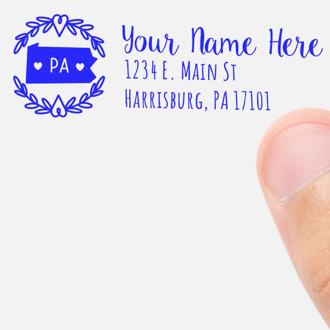 Pennsylvania State Custom Return Address Stamp in use, featuring a blue state outline with hearts and text for name and address. A thumb is visible, holding the paper.