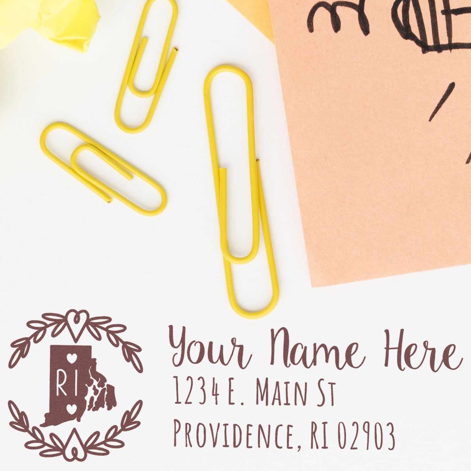 Yellow paperclips and a brown envelope beside a PSI Pre-Inked Rhode Island State Customized Address Stamp impression with a decorative border, showing a sample address in Providence, RI.