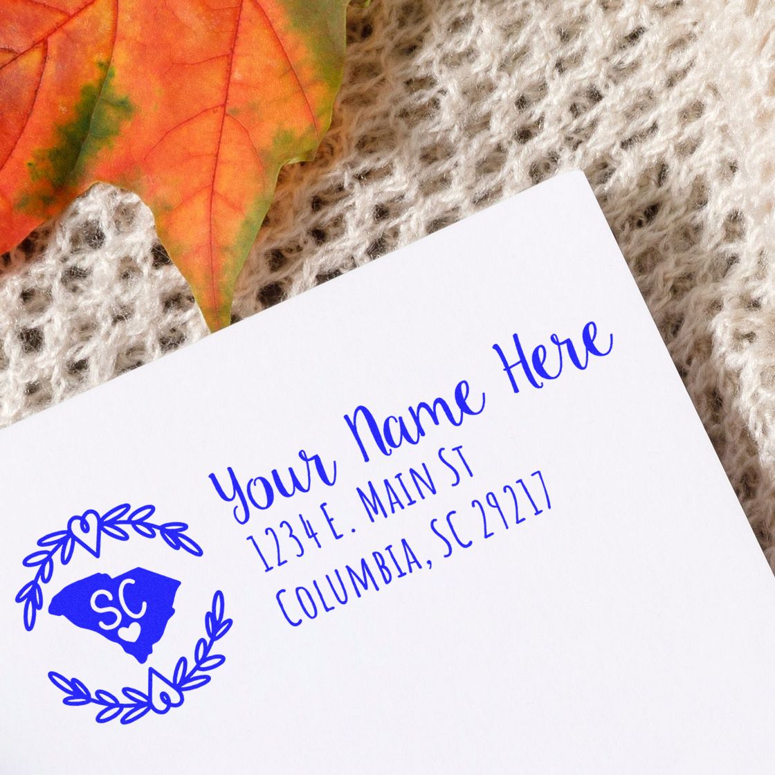 PSI Pre-Inked South Carolina State Customized Address Stamp on a white envelope with a blue imprint, featuring a heart and state outline, placed on a knitted surface with a fall leaf nearby.