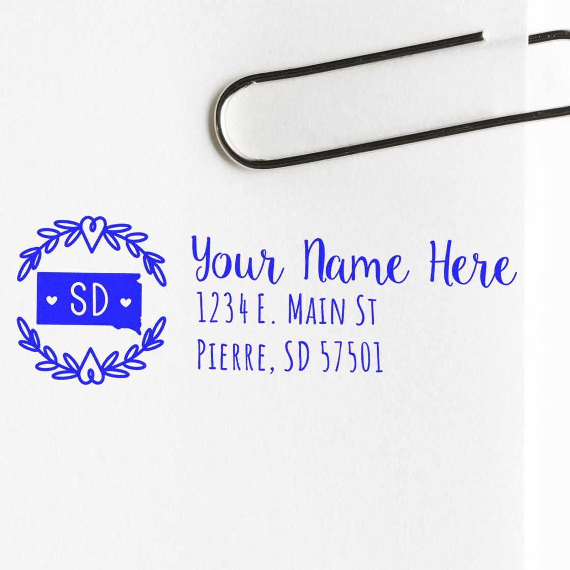 South Dakota State Custom Return Address Stamp in blue ink on white paper, featuring a state outline with SD and hearts, next to a sample address. A paperclip is visible at the top.