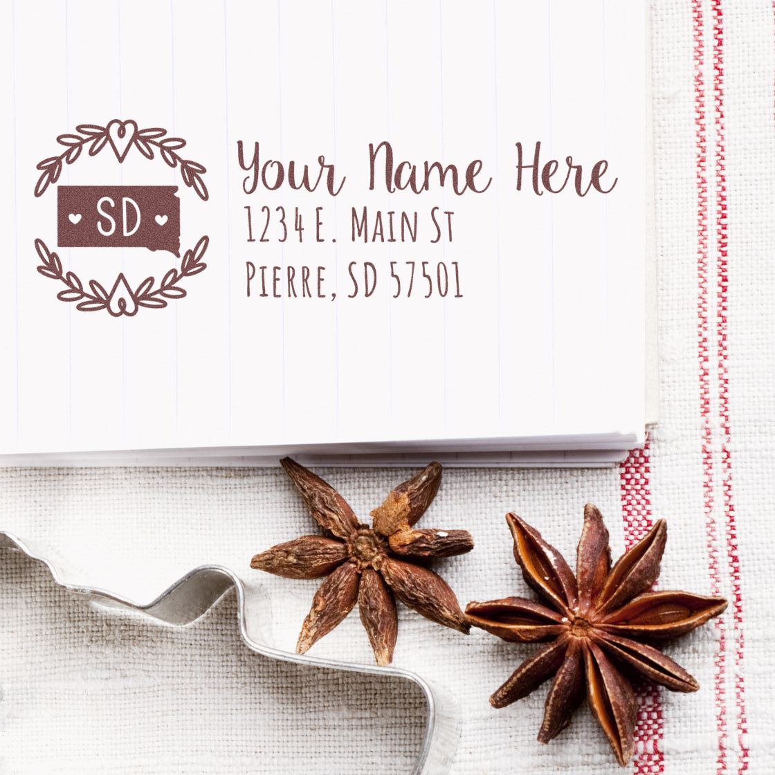 Image of a South Dakota State Custom Return Address Stamp on paper with a decorative border, featuring placeholder text for name and address. Star anise and a cookie cutter are placed nearby.