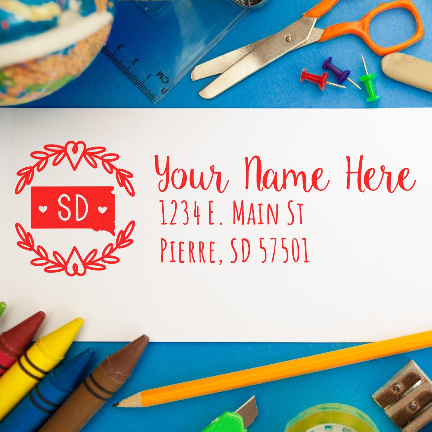 South Dakota State Custom Return Address Stamp on an envelope surrounded by colorful stationery, including scissors, pencils, and push pins. The stamp features a heart design and state outline.