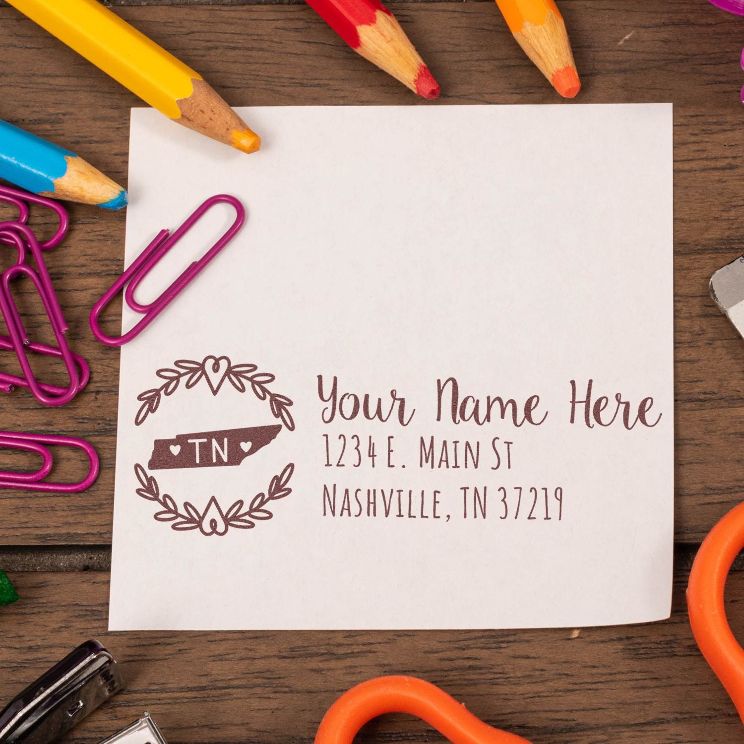 A Self-Inking State Wreath of Tennessee Address Stamp on a white paper with a decorative wreath design, surrounded by colorful pencils and paperclips on a wooden desk.