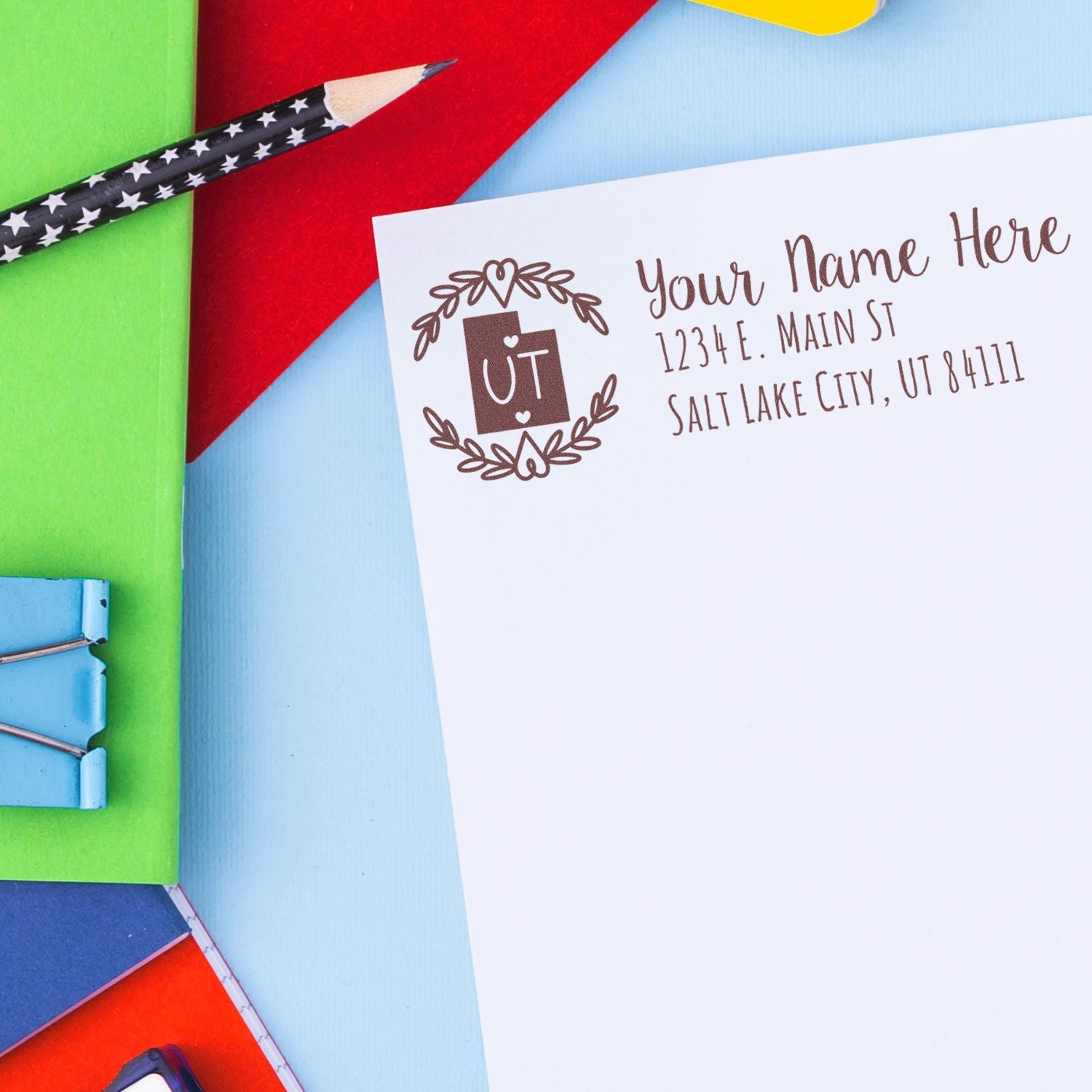 Utah State Custom Return Address Stamp on a white envelope, featuring a decorative UT design. Surrounded by colorful stationery, including a star-patterned pencil and paper clips.