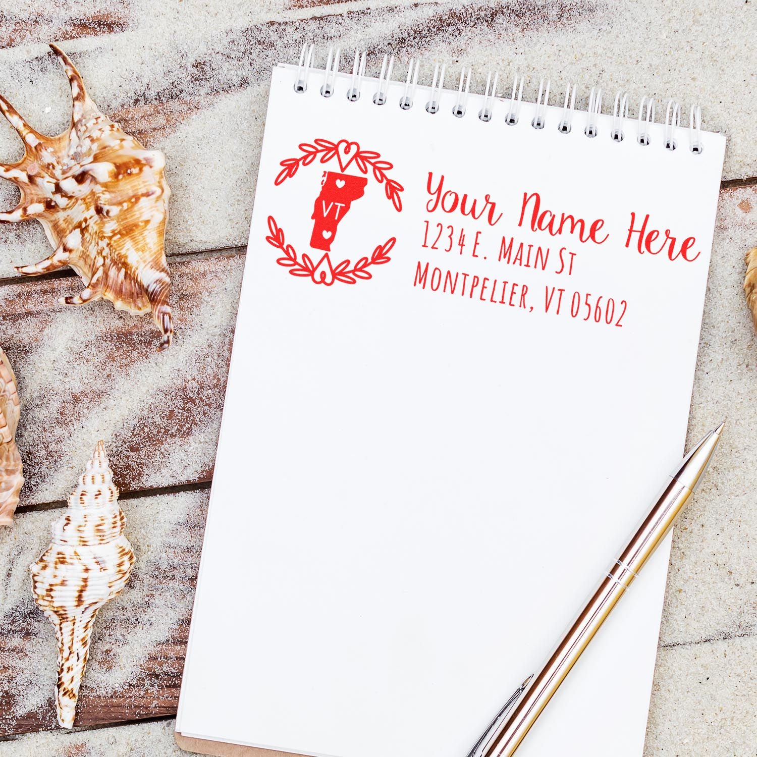 Notebook on sand with seashells, featuring a Vermont State Custom Return Address Stamp design in red, displaying placeholder text for name and address. Pen placed beside the notebook.