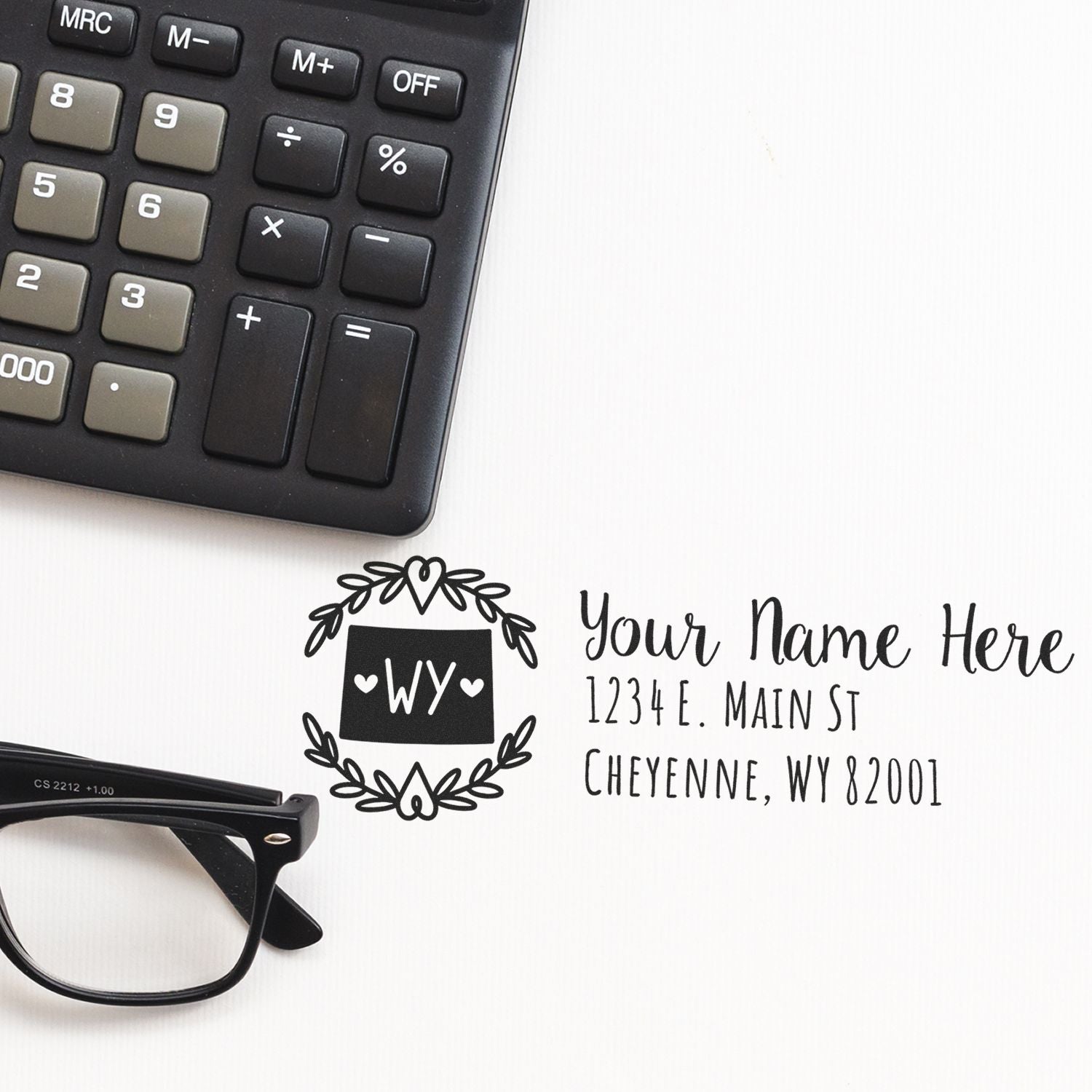 Self-Inking State Wreath of Wyoming Address Stamp on a desk with a calculator and glasses, featuring a decorative wreath design and customizable address text.