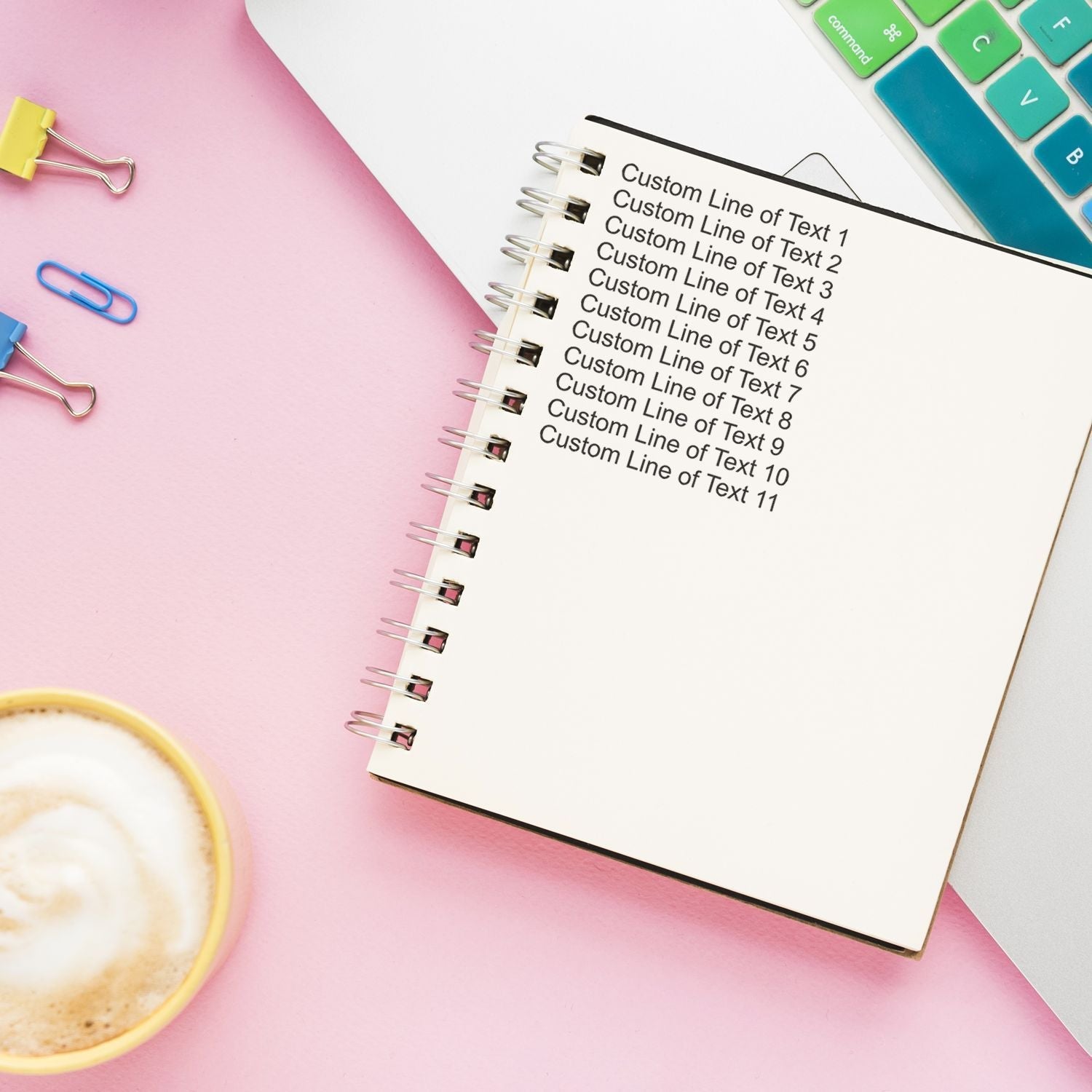 11 Line Custom Rubber Stamp with Wood Handle used on a notebook, placed on a pink desk with a coffee cup, paper clips, and a colorful keyboard.