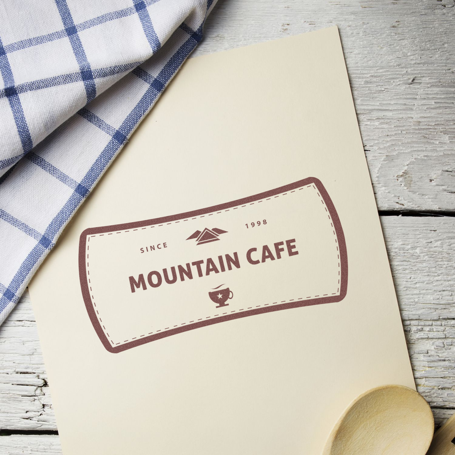 Custom Rubber Stamp Size 3 x 7 with Mountain Cafe logo on paper, placed on a wooden surface with a checkered cloth and wooden spoon.