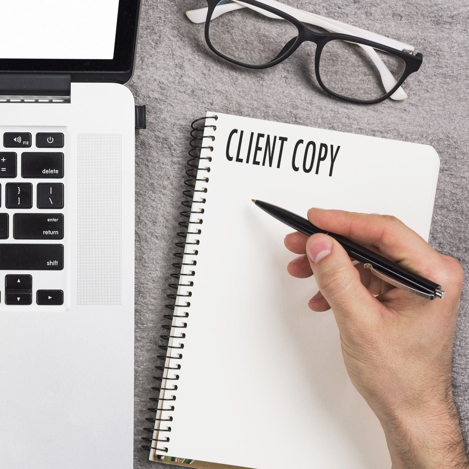 A hand holding a pen next to a Large Pre-Inked Client Copy Stamp, with CLIENT COPY stamped on a notebook, beside a laptop and glasses.