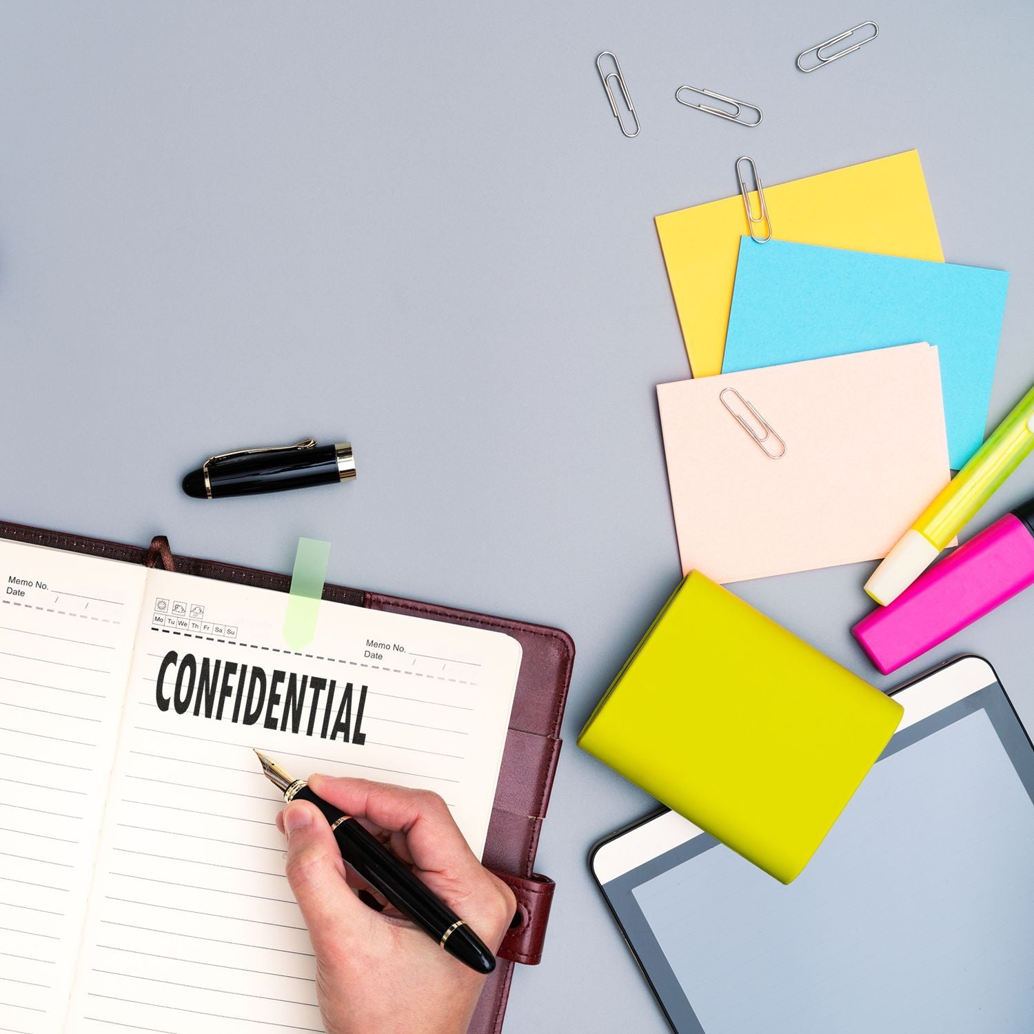 Person using a Large Pre-Inked Confidential Stamp on a notebook, surrounded by colorful sticky notes, paper clips, a pen, and a tablet.