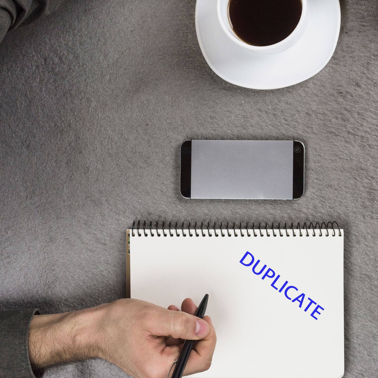 A hand holding a pen near a notebook stamped with DUPLICATE using a Large Pre-Inked Duplicate Stamp, with a phone and coffee cup nearby.