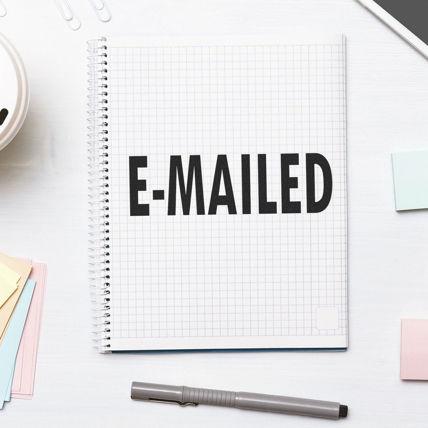 Large Pre-Inked E-Mailed Stamp on a grid notebook, surrounded by sticky notes, a pen, and office supplies on a white desk.