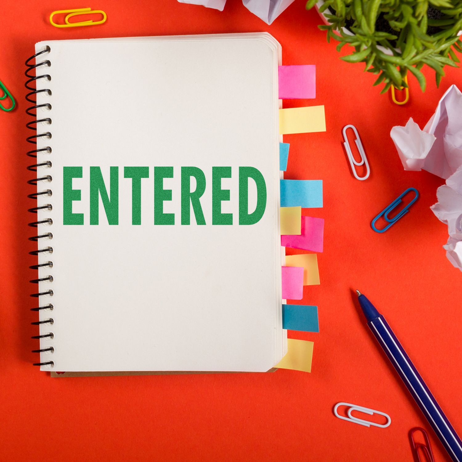 Notebook stamped with ENTERED using the Large Pre-Inked Entered Stamp, surrounded by colorful paper clips, sticky notes, and a pen on a red background.