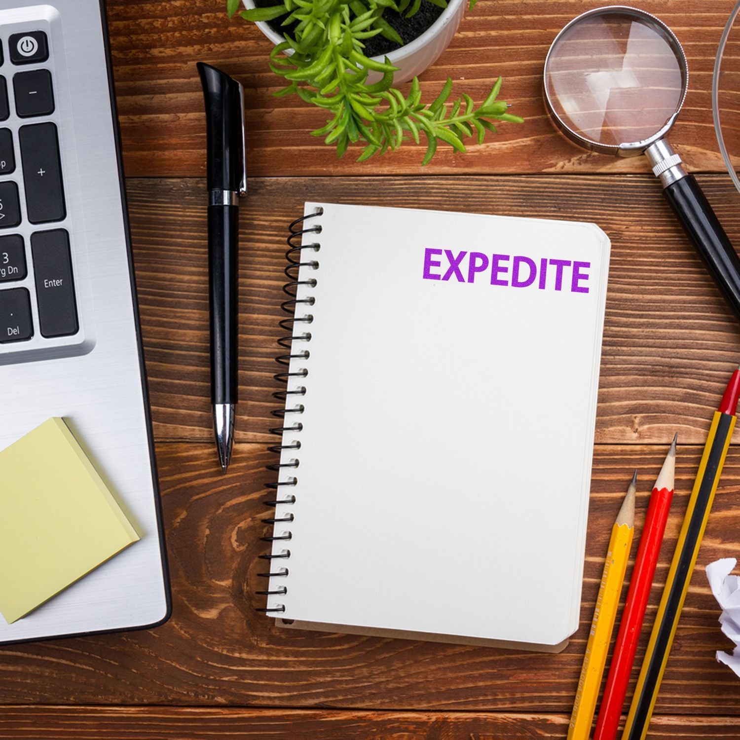 A desk with a laptop, pen, sticky notes, magnifying glass, pencils, plant, and a notebook stamped with EXPEDITE using the Large Pre-Inked Expedite Stamp.